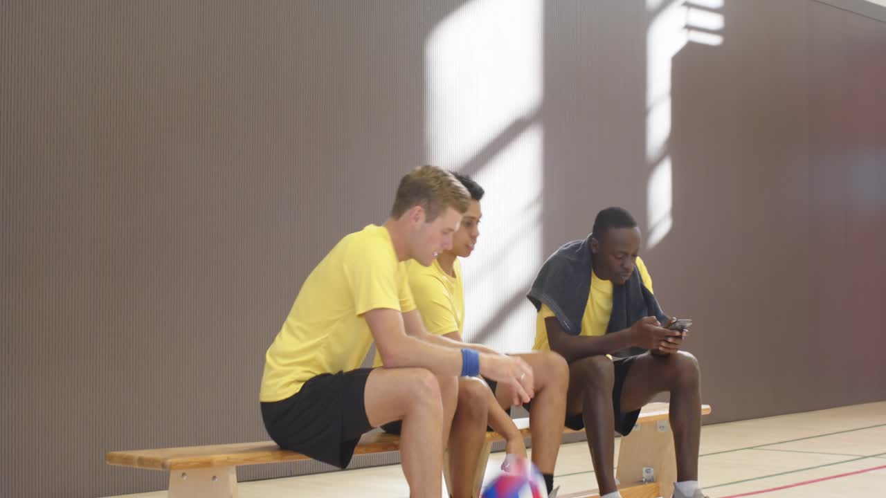 Diverse men in yellow entering gym sitting bench checking phone holding volleyball and bottle preppi