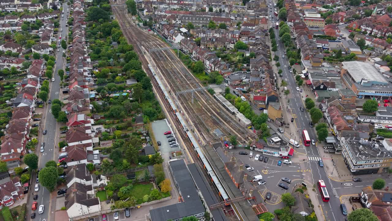 Slow aerial pan across Chingford station with eight train tracks, a stationary train, light road traffic, buses, cars, and nearby houses and offices, capturing the wider urban transport landscape