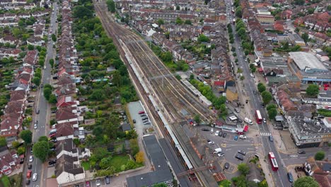 slow aerial pan across chingford station with eight train tracks, a stationary train, light road traffic, buses, cars, and nearby houses and offices, capturing the wider urban transport landscape