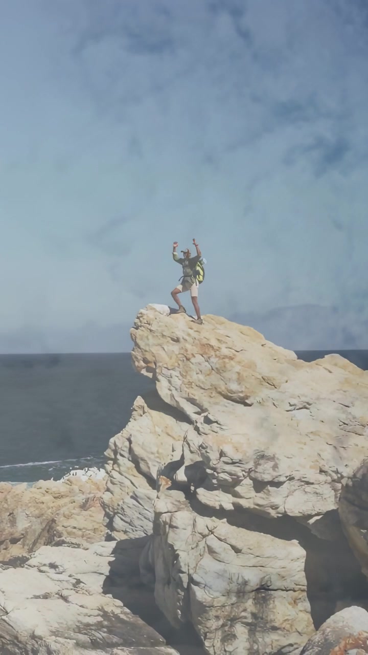 Escursionista uomo che raggiunge la vetta di una roccia e alza le braccia, celebrando la conquista con l'orizzonte dell'oceano