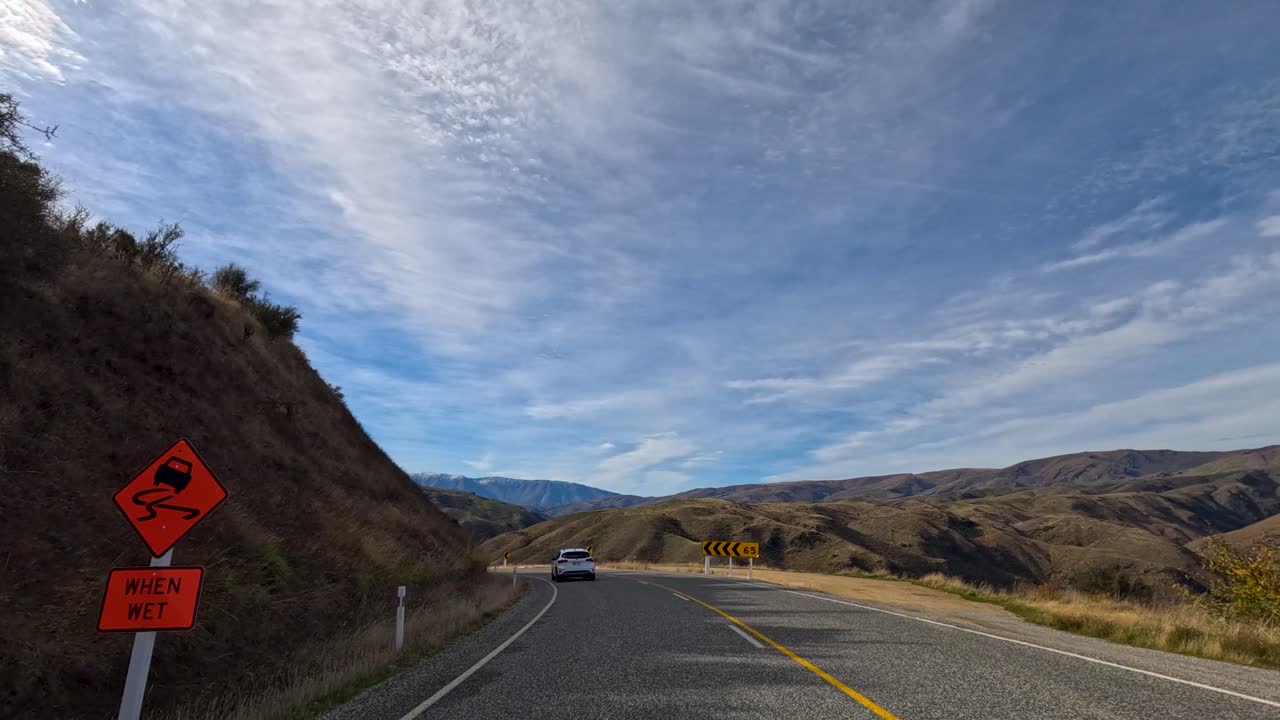 A car travels through winding roads in New Zealand, surrounded by autumn hills under a bright, partly cloudy sky