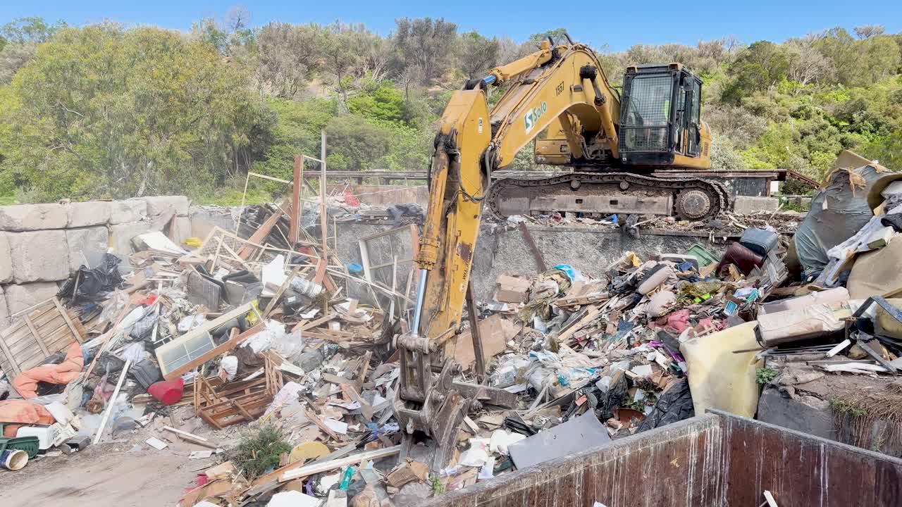 Excavator lifts and moves rubbish in a landfill surrounded by trees and blue sky