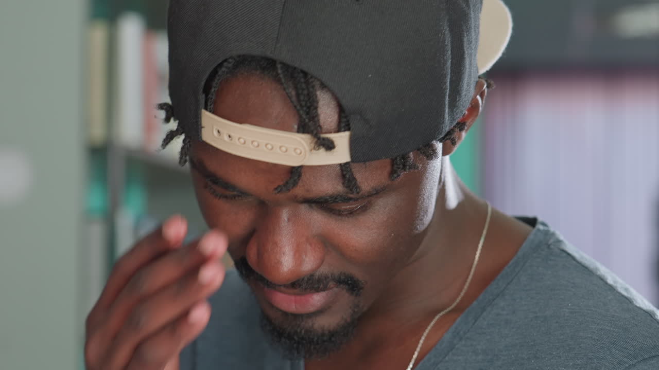 Close up of young man wearing backward cap smiling softly while tilting head down, showing relaxed expression and subtle dimples, in warm indoor setting with soft background