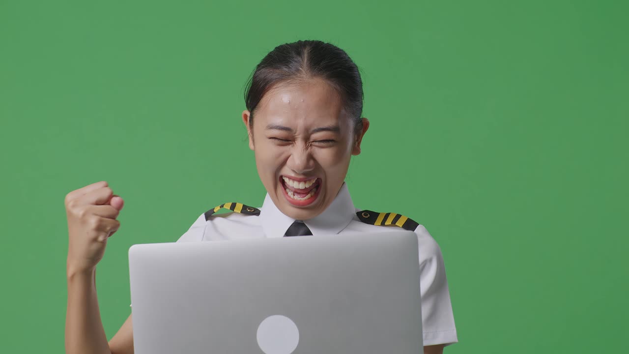 아시아 여성 조종사가 노트북을 바라보고 그 다음 녹색 스크린 배경 스튜디오에 서서 축하하는 골을 비명을 지르고 있습니다.