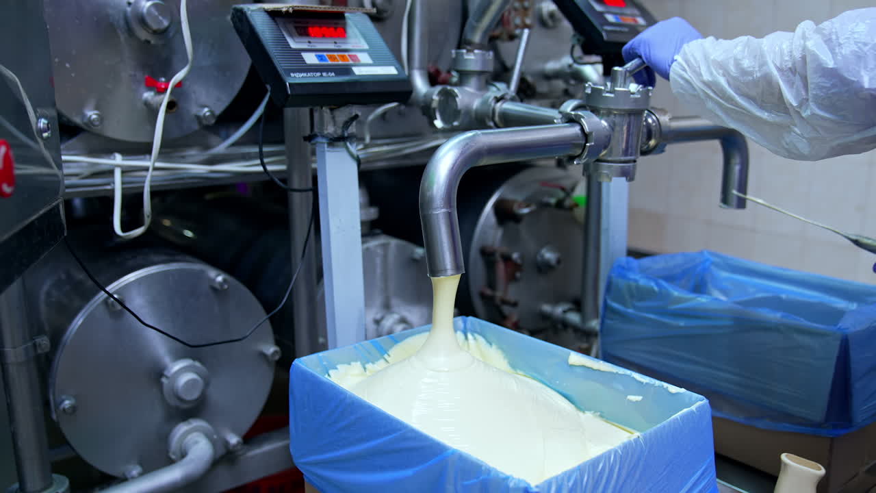 Creamy white dairy substance being poured into the box covered with plastic bag. Hand of the worker pushes the valve to stop pouring. Filling another box with dairy product.