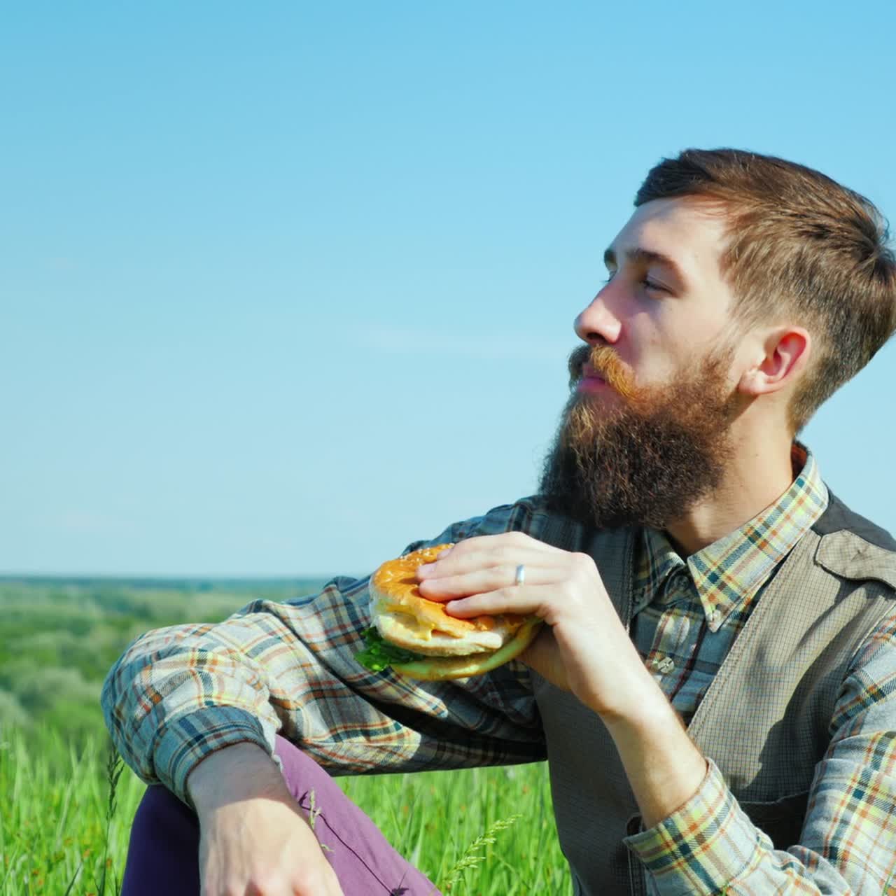 un joven barbudo comiendo una hamburguesa al aire libre