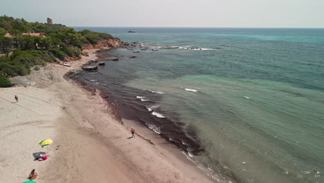 Blue-Sea-And-Beach-With-Tourists-Enjoying-The-Summer-Holidays-In-Sardinia,-Italy---aerial-pullback