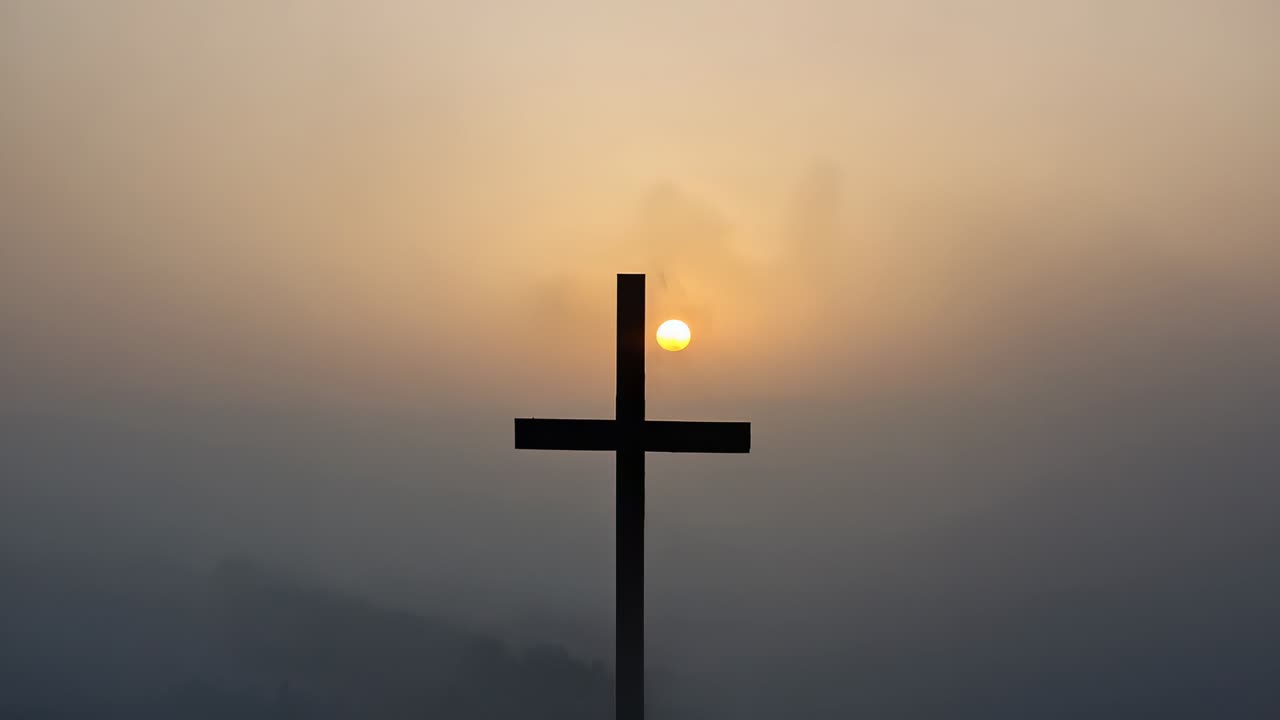 Sun peeking behind metal cross and climbing above crossbar over foggy hilltop, illuminating mist