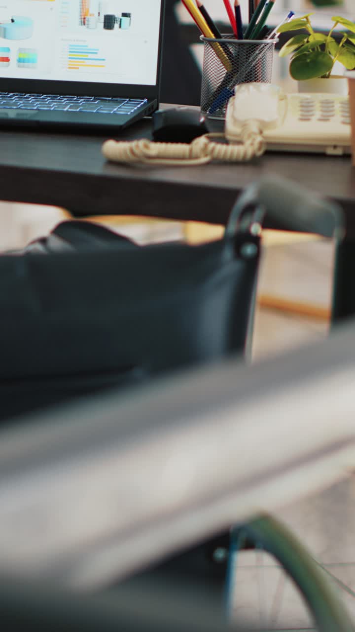 Vertikales Video: Rollstuhl im Büro und Laptop auf dem Schreibtisch zeigt Geschäftsanalysen.