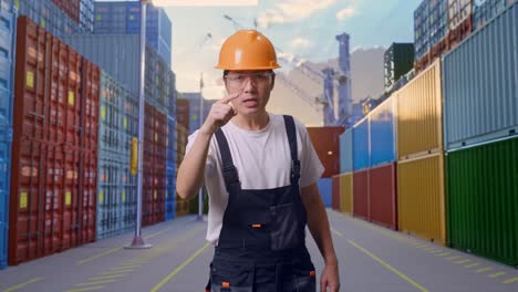 trabajador asiático con gafas de protección y casco de seguridad gritando a la cámara mientras está de pie en el almacén del patio de contenedores