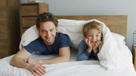 cheerful funny father and little son laughing and looking out of the blanket while lying on the bed in the morning