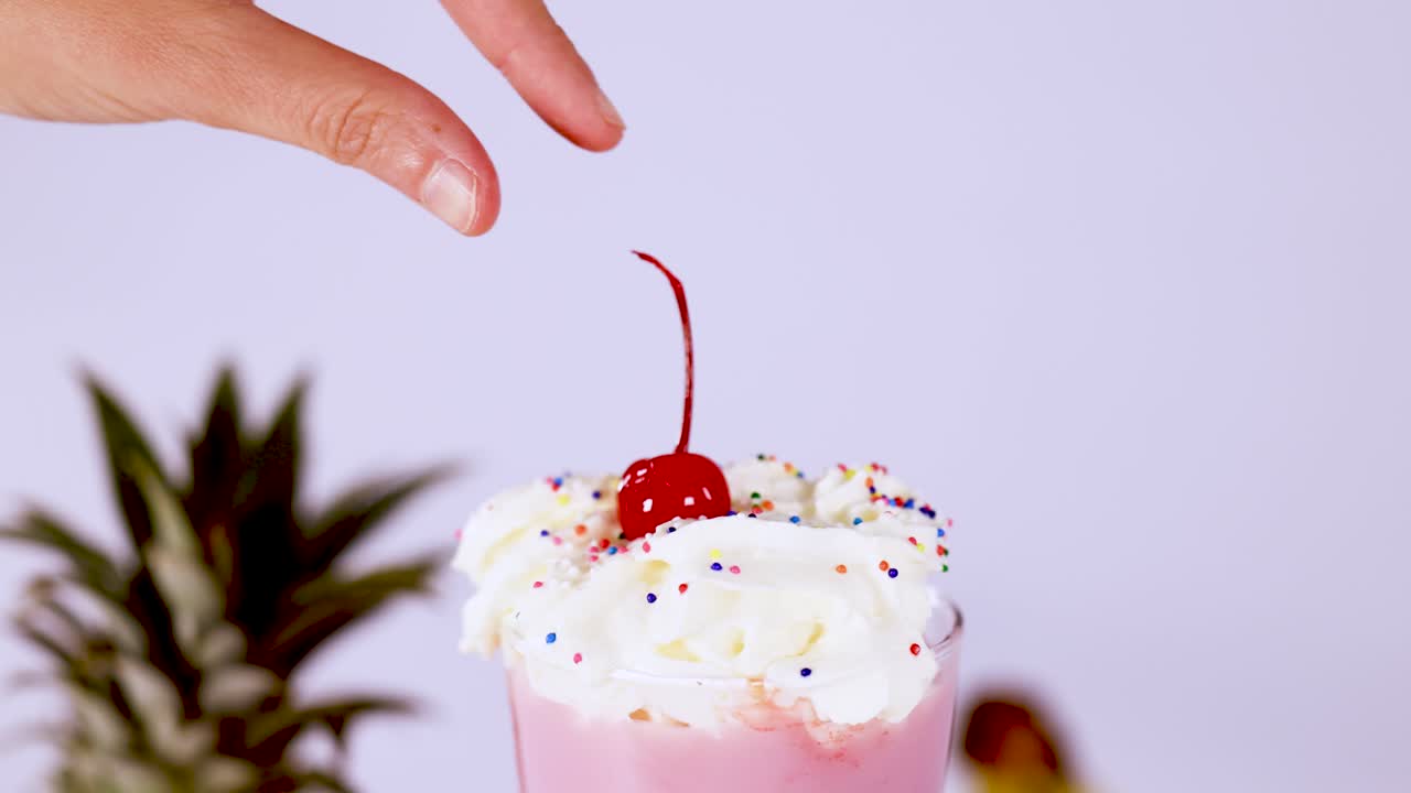Hand removes cherry from pink milkshake topped with whipped cream and sprinkles, bright lighting