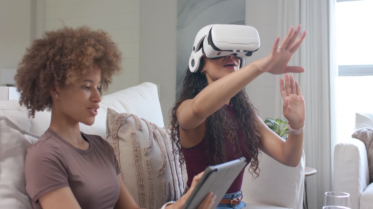 African American women friends on sofa right putting VR headset interacting, left tapping tablet