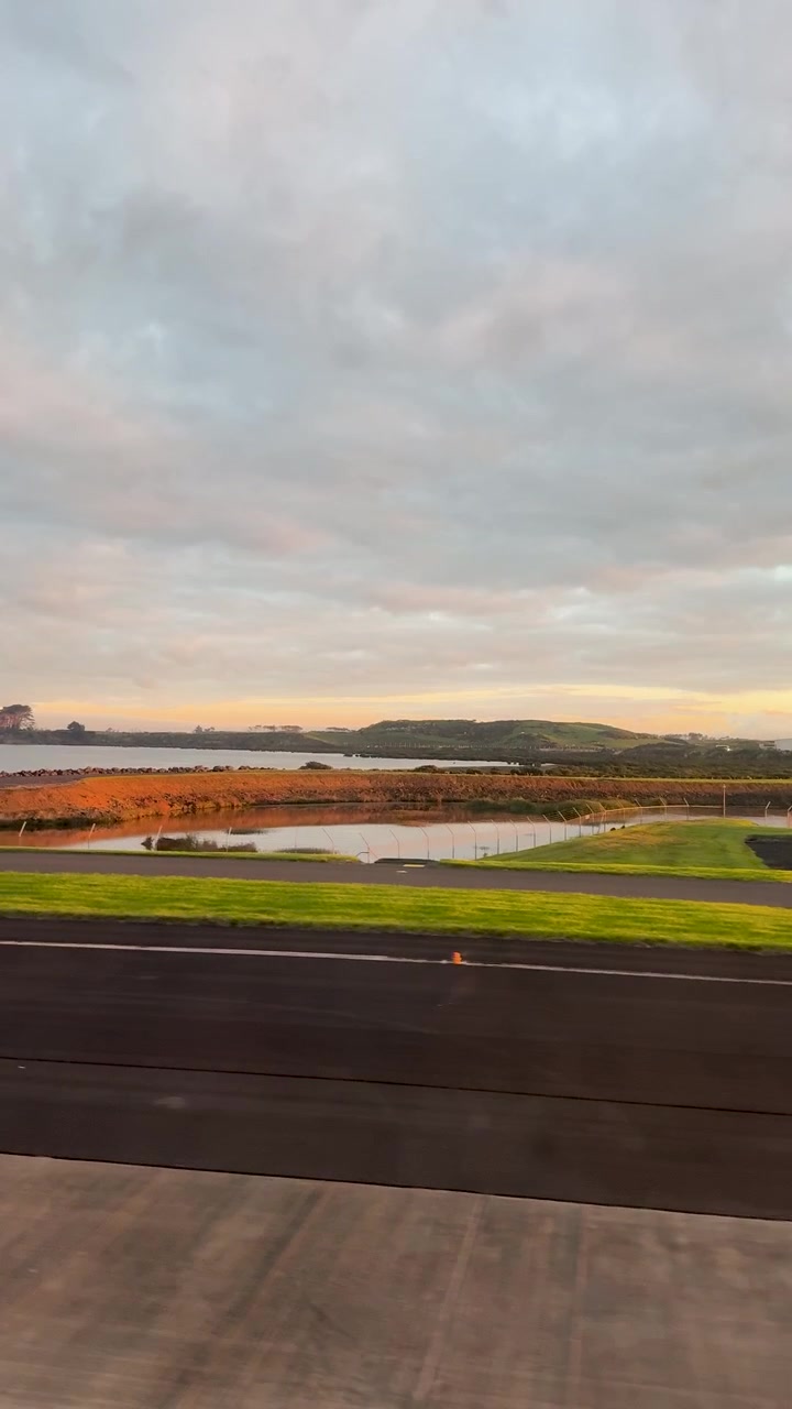 Pesawat lepas landas dari Bandara Auckland, menampilkan pemandangan landasan pacu dan lanskap di bawah cahaya fajar yang lembut.