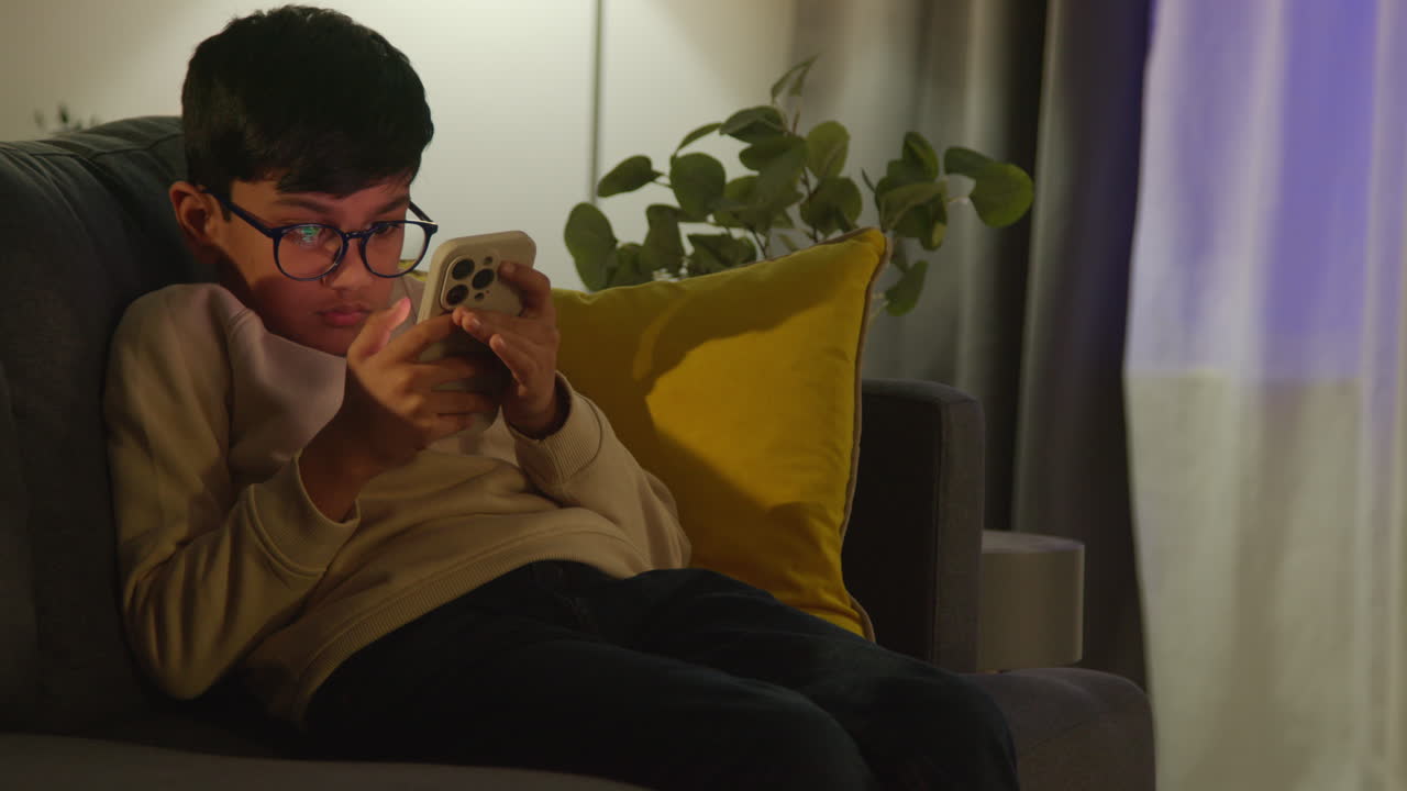 어린 소년이 집에서 휴게실에서 소파에 앉아서 밤에 드폰으로 게임을 하고 있다.