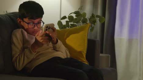 young boy sitting on sofa in lounge at home playing game on mobile phone at night 1