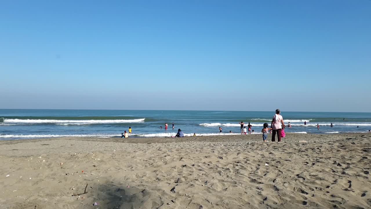 вид сзади на бабушку и молодую девушку, идущих к берегу с людьми, как детьми, так и взрослыми, наслаждающимися волнами у береговой линии общественного пляжа