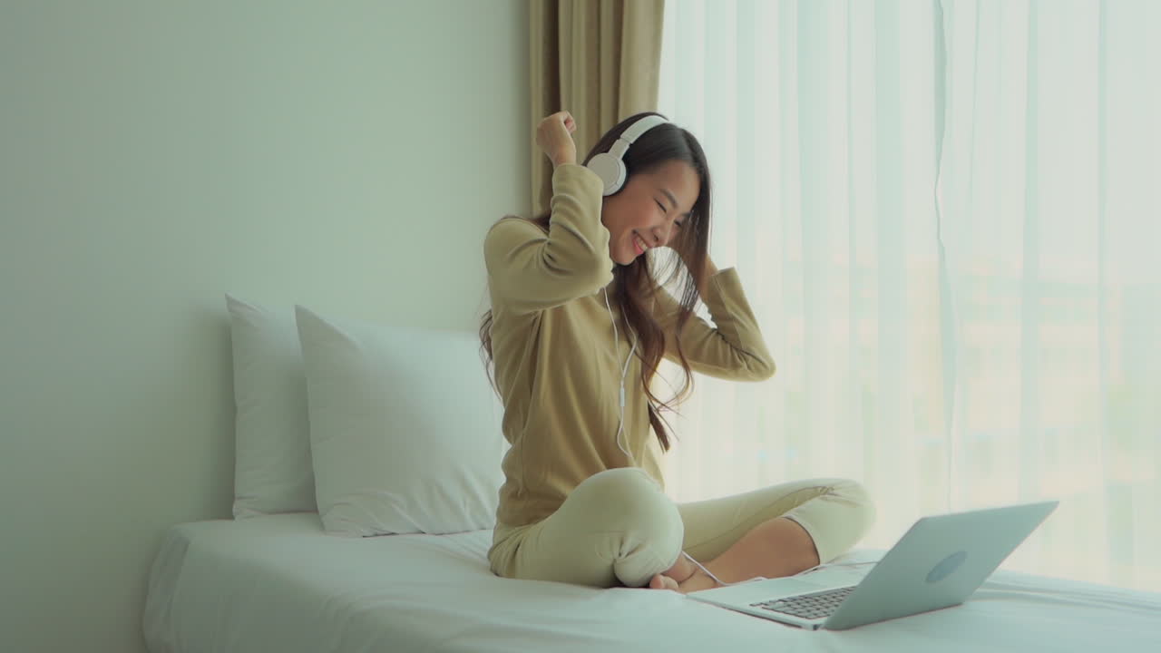 A young, Asian woman is dancing to the music playing from her laptop while sitting on the bed