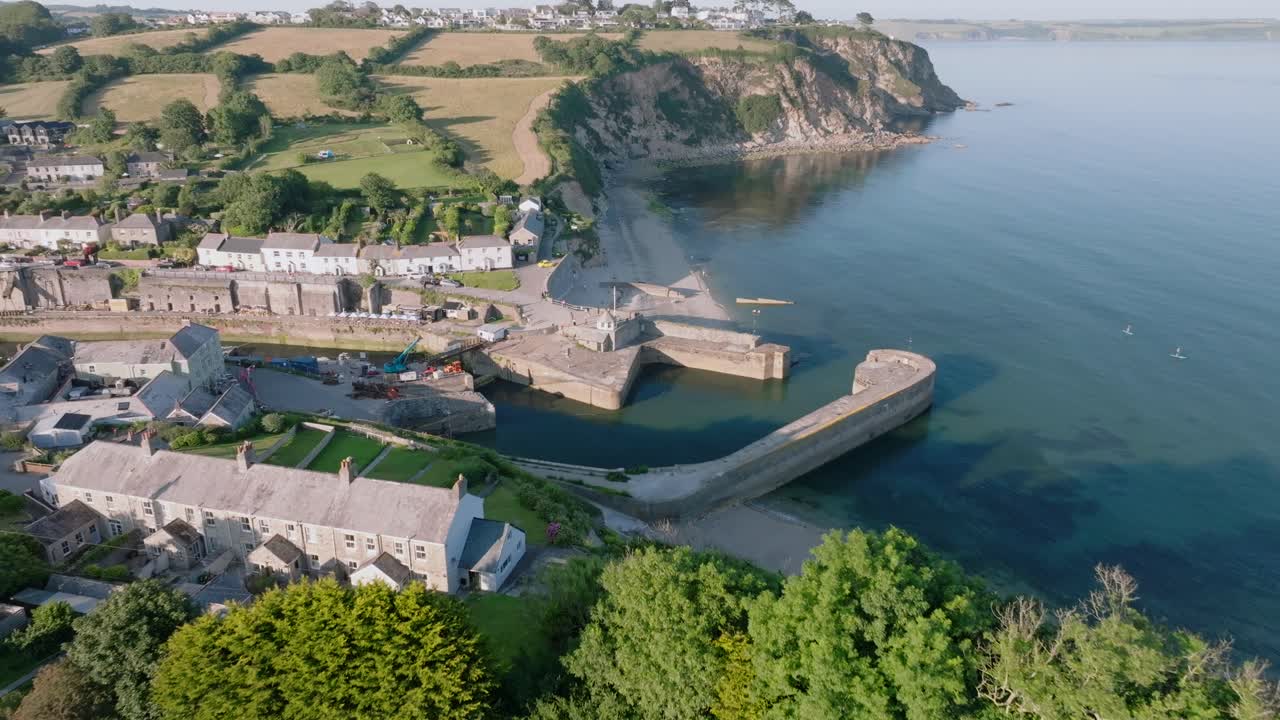 Harbour village with camera flying over green fields and trees towards port opening and sea cliffs. Charlestown, Cornwall, UK.