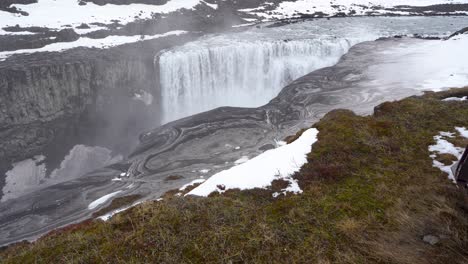 una vista impresionante de una majestuosa cascada en islandia con patrones giratorios en el río debajo, manchas de nieve y cielos nublados