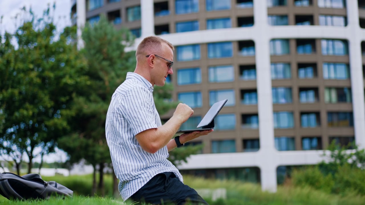 Man using a tablet with a keyboard outdoors. A seated man in sunglasses holds a device beside grass, with apartment windows in the background.