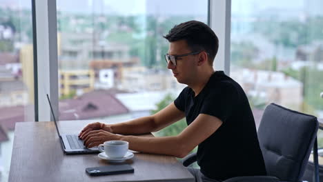 young adult office employee working at laptop. dark-haired man focused on work. blurred cityscape in window at backdrop.