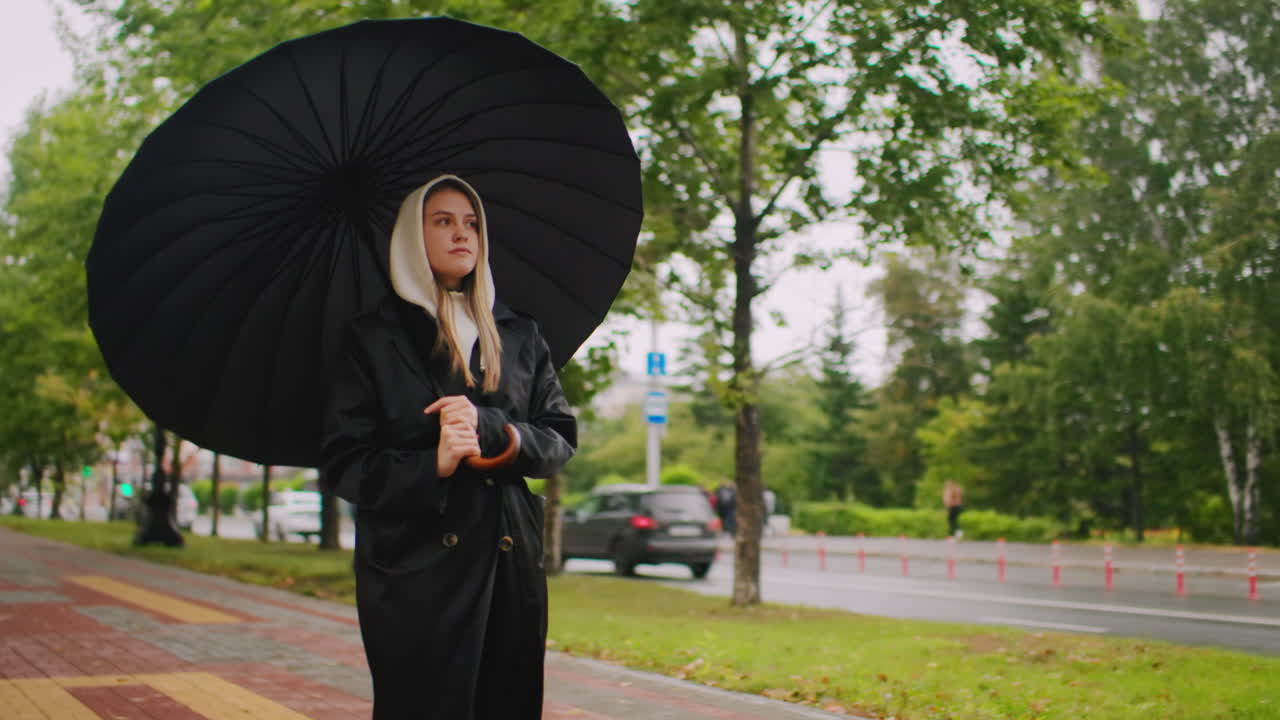 Young woman in long black coat and hood holding umbrella walking along tree-lined pathway with fallen leaves, cars and shops in background, calm urban lifestyle moment during rainy autumn day