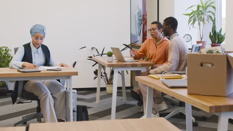 Muslim-Businesswoman-Sitting-At-A-Table-Taking-Notes-While-An-Businesswoman-Talks-To-The-New-Employee-Looking-At-Laptop-At-Another-Table-In-The-Office-2