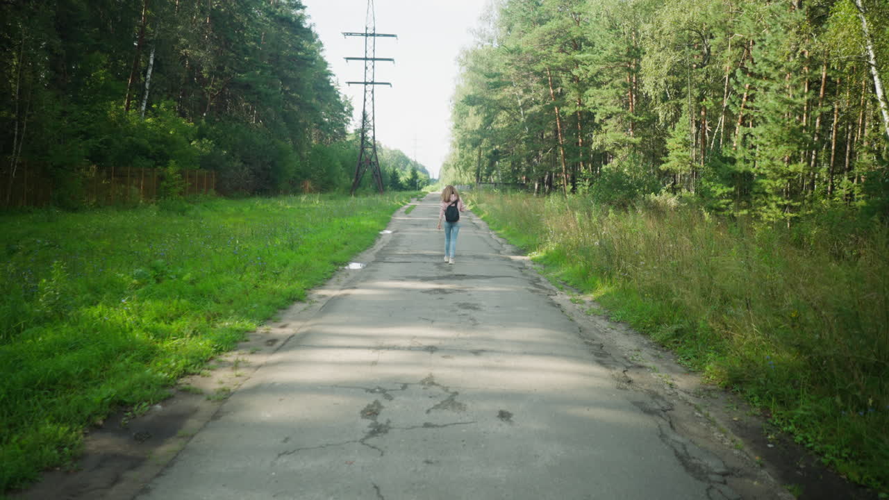 Distant view of lady walking alone on tarred country road lined with dense green forest, overhead power poles, and scattered puddles under daylight, creating a calm and introspective atmosphere