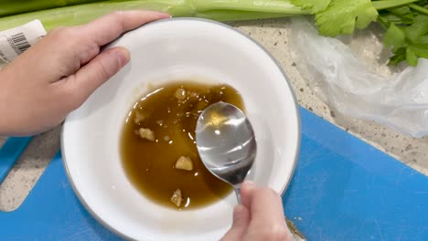 hands use a spoon to mix fish sauce, sugar, and chopped garlic in a white bowl on a kitchen counter with natural lighting and overhead view