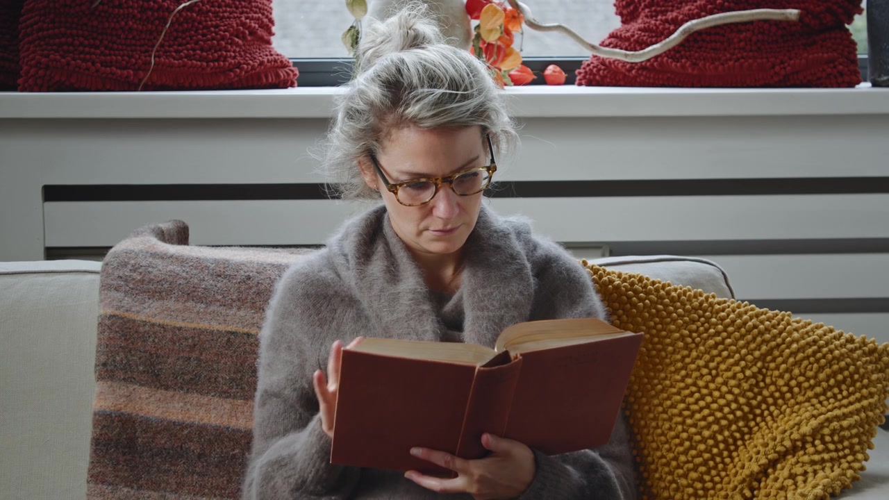 Vue d'ensemble d'une jeune femme caucasienne à lunettes assise sur un canapé, lisant et étudiant intensément dans le salon moderne d'un appartement.