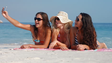 happy pretty friends lying on towel and taking a selfie at the beach