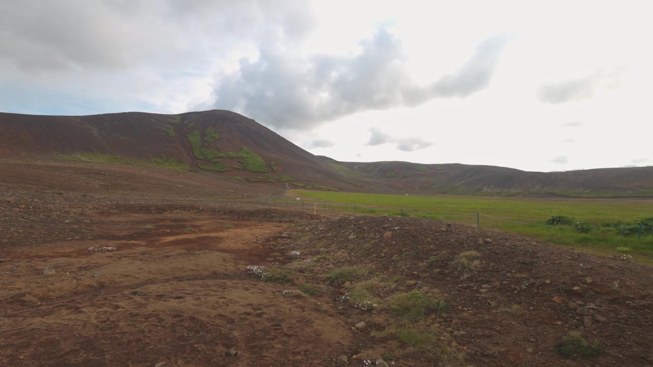 Sebuah bidikan lebar sinematik dari medan vulkanik terjal di Islandia, menampilkan tanah berbatu dan pegunungan yang jauh. Menangkap keindahan alam Nordik yang otentik dan minimalis