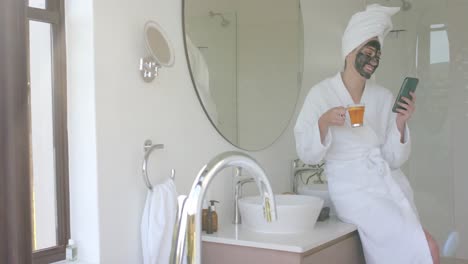 mujer caucásica feliz con máscara bebiendo té y usando teléfono inteligente en el baño en cámara lenta