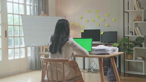 vista trasera de una mujer asiática hablando con una computadora portátil de pantalla verde mientras trabaja con documentos en la oficina