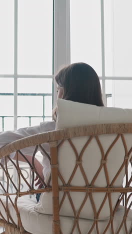 brunette young woman artist rests after work sitting in rattan armchair with pillows near easel with picture in studio backside view slow motion
