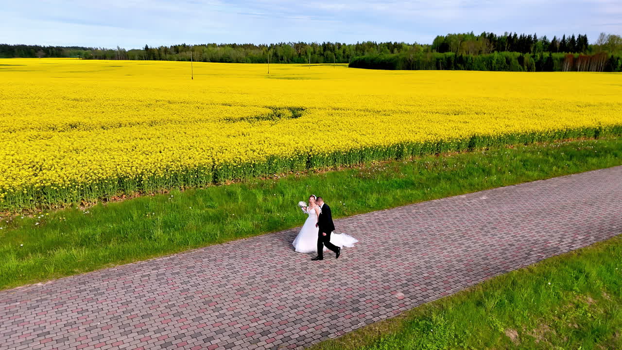 A bride and groom walk hand in hand along a paved country road next to a vibrant yellow flower field
