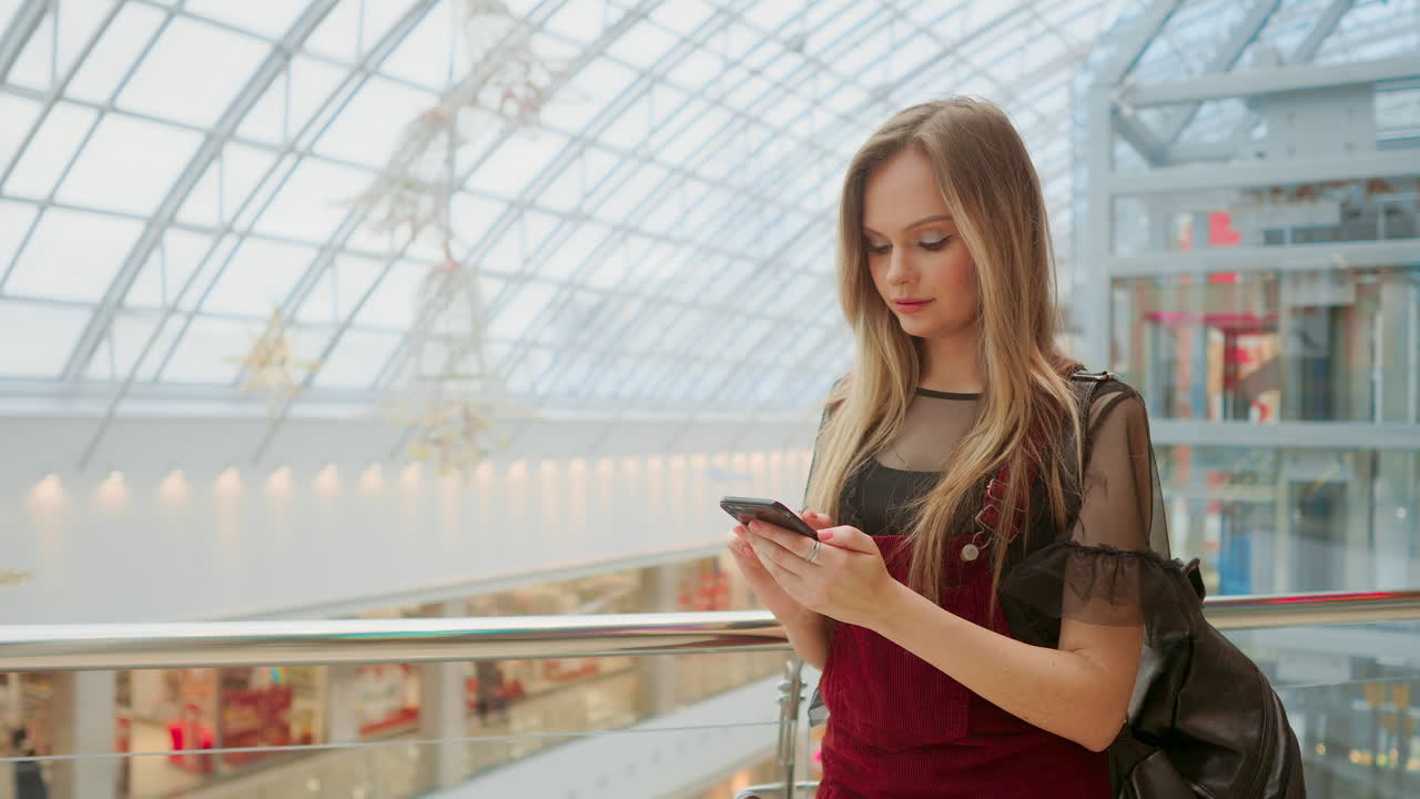 Young tourist woman in hat, with backpack stands at airport and uses smartphone.Hipster girl checks email, chatting, blogging, browsing internet.Online marketing, education. Distance work, e-learning.