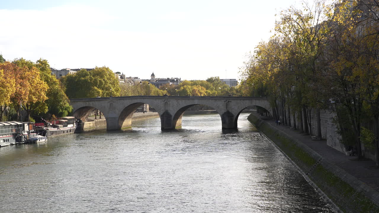 Sunny autumn view of Pont Marie over the Seine River in Paris, with golden trees lining the banks, calm water, and moored boats along the riverside under soft afternoon light