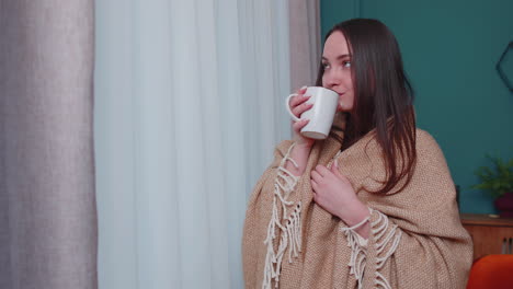 Young-adult-brunette-girl-in-warm-white-sweater-drink-hot-beverage-mug-relaxing-at-home-near-window