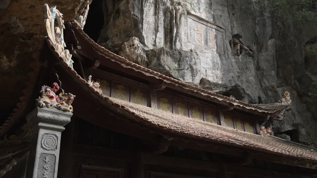 Ornate tiled roof of Bich Dong Pagoda beneath a weathered limestone cliff with carved reliefs and cave niches in Ninh Binh, Vietnam, finished with a calm upward tilt handheld shot