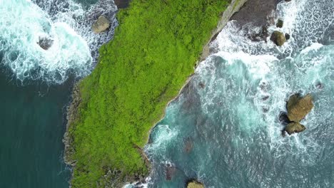 aerial top down shot of green island with crashing waves against rocky cliffs