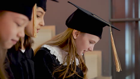 cerca de tres niñas felices con toga y birrete hablando juntas mientras se sientan en las escaleras en la ceremonia de graduación de preescolar