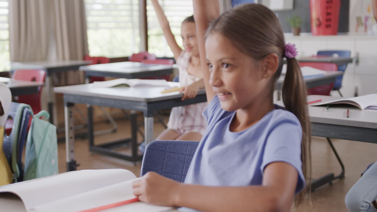 alumnas felices y diversas en los escritorios levantando las manos en el aula de la escuela primaria