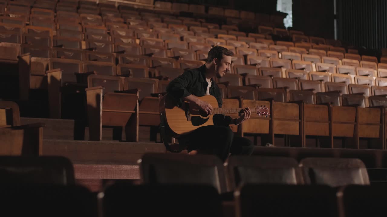 un chico guapo toca una guitarra acústica en un cine abandonado. el músico canta una canción y acompaña en la guitarra