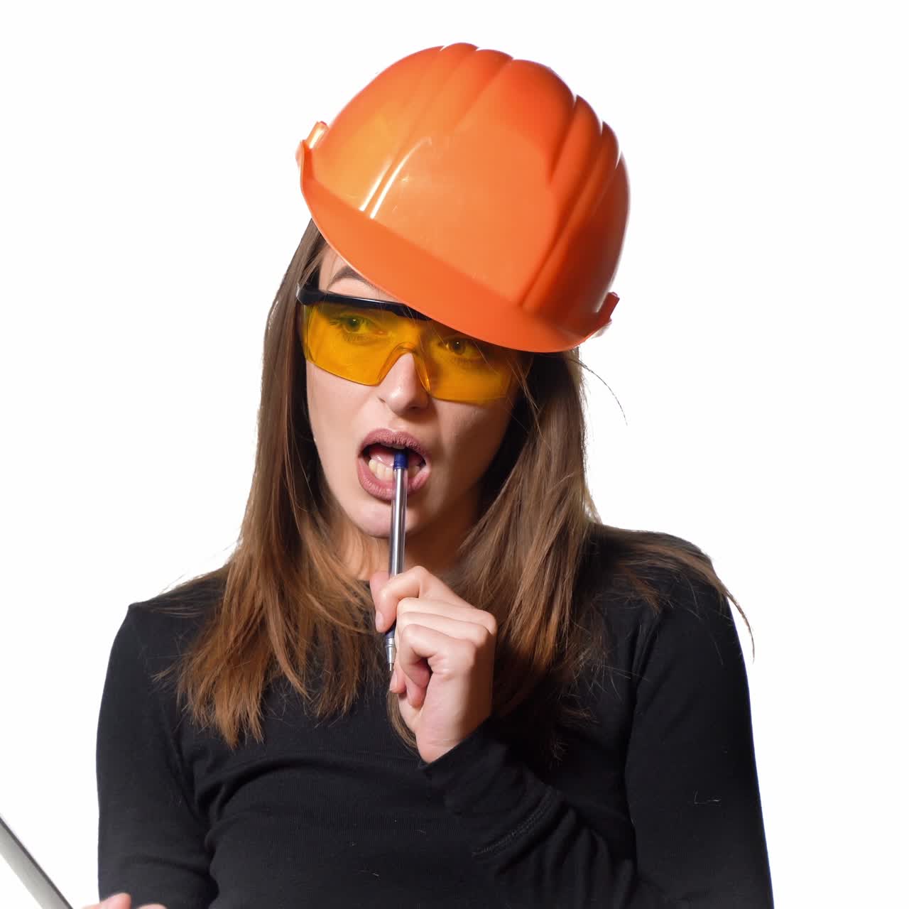 A woman in an orange helmet and protective eyeglasses is talking and holding a pen and folder in her hands on a white background. Features of work of the engineer