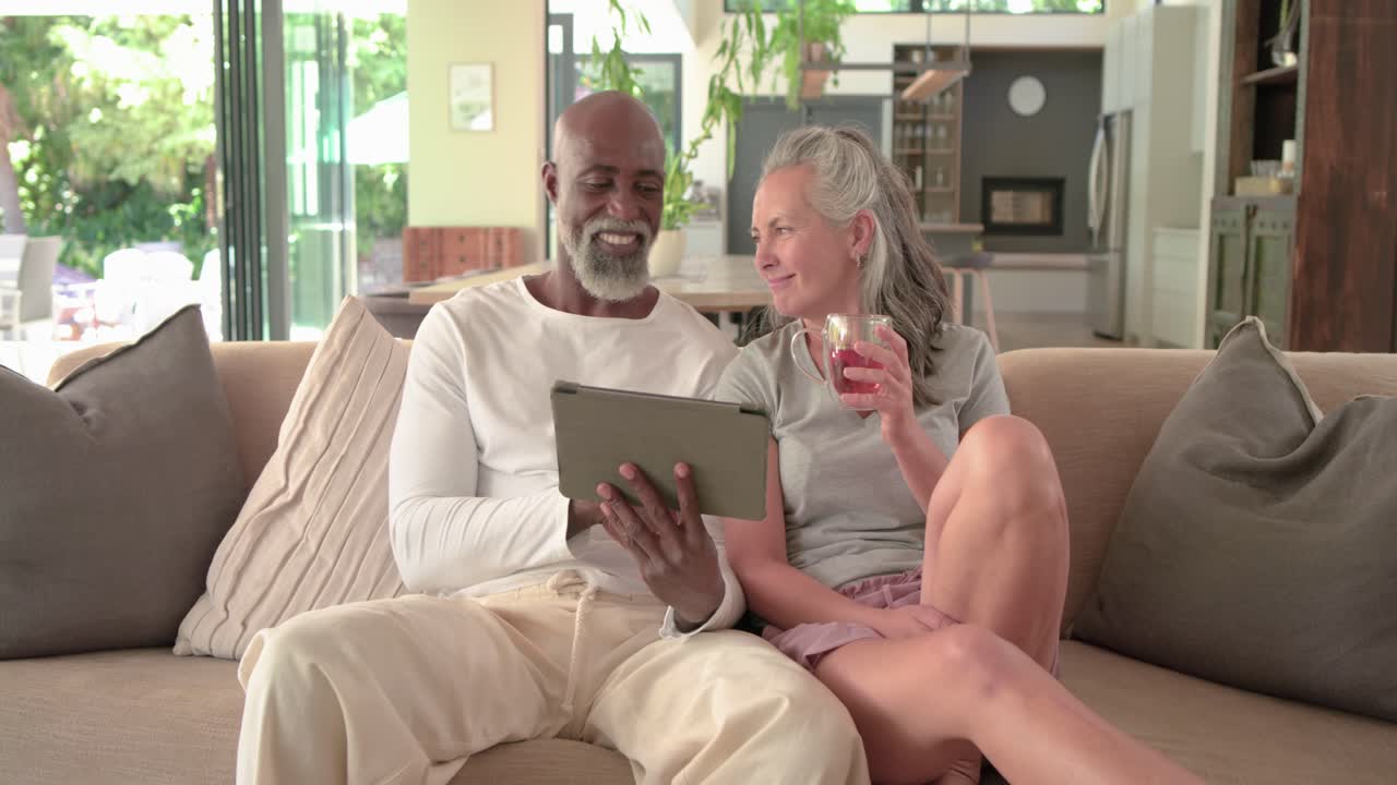 Coppia multietnica seduta sul divano a casa, l'uomo inclina il tablet condividendo contenuti, la donna sorseggia una bevanda rossa