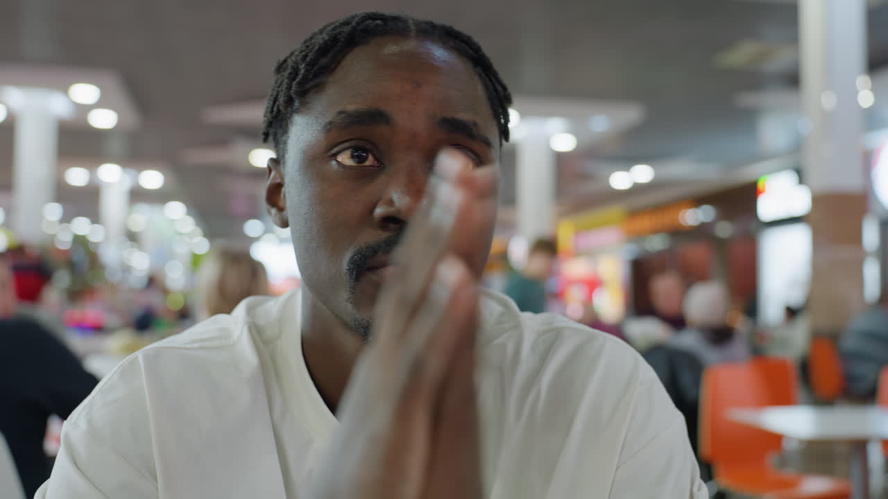 Young african man in white shirt sits in busy restaurant rubbing face with both palms in tired or stressed mood while people interact in background under bright indoor lighting