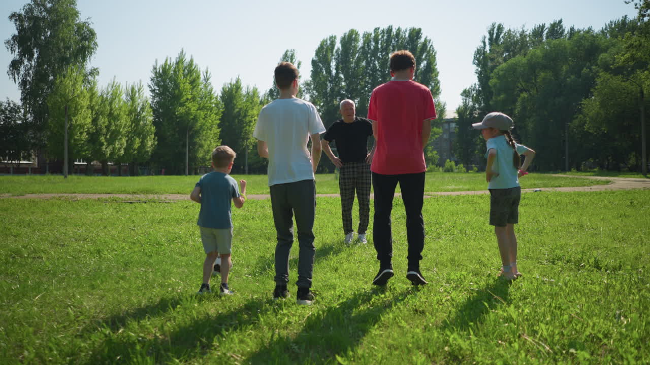 un abuelo conduce a sus nietos en un ejercicio de salto en un campo cubierto de hierba, se paran juntos, manos en la cintura, y saltan al unísono. una pelota se encuentra cerca