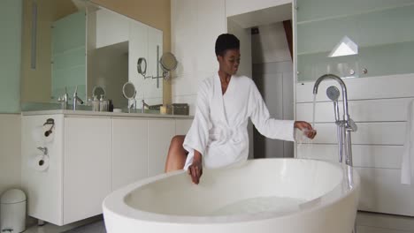 Smiling-african-american-attractive-woman-in-white-robe-sitting-on-bathtub,-preparing-bath